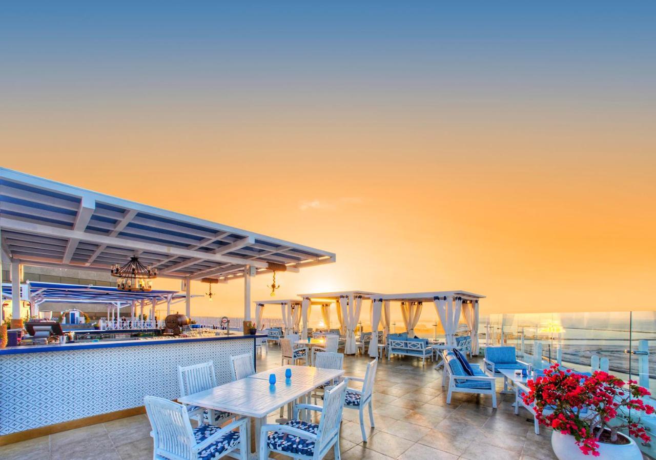 Hyatt Regency Dubai - Corniche Hotel Екстериор снимка The photo depicts a stylish rooftop terrace with a vibrant sunset in the background. The scene includes a bar area with white furnishings and blue accents, as well as several lounge chairs. There are elegant cabanas draped nearby, and colorful flower